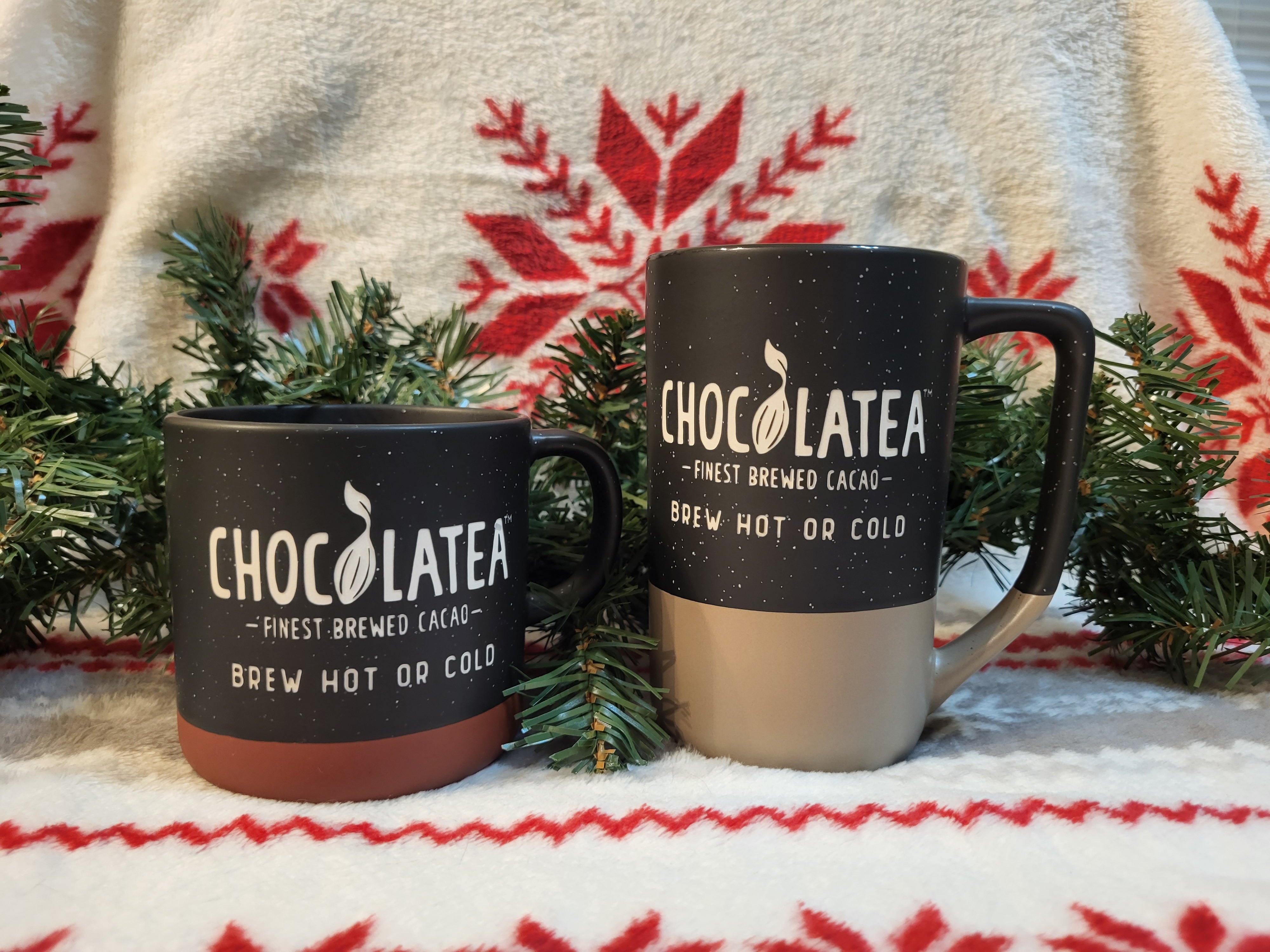 Two mugs labeled "CHOCOLATEA," one black and one brown-and-tan, sit among greenery with a festive blanket backdrop, featuring Dark Chocolate Mocha.