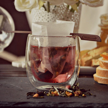 A glass cup of vibrant organic tea with a steeping bag, on slate beside bread slices and flowers.