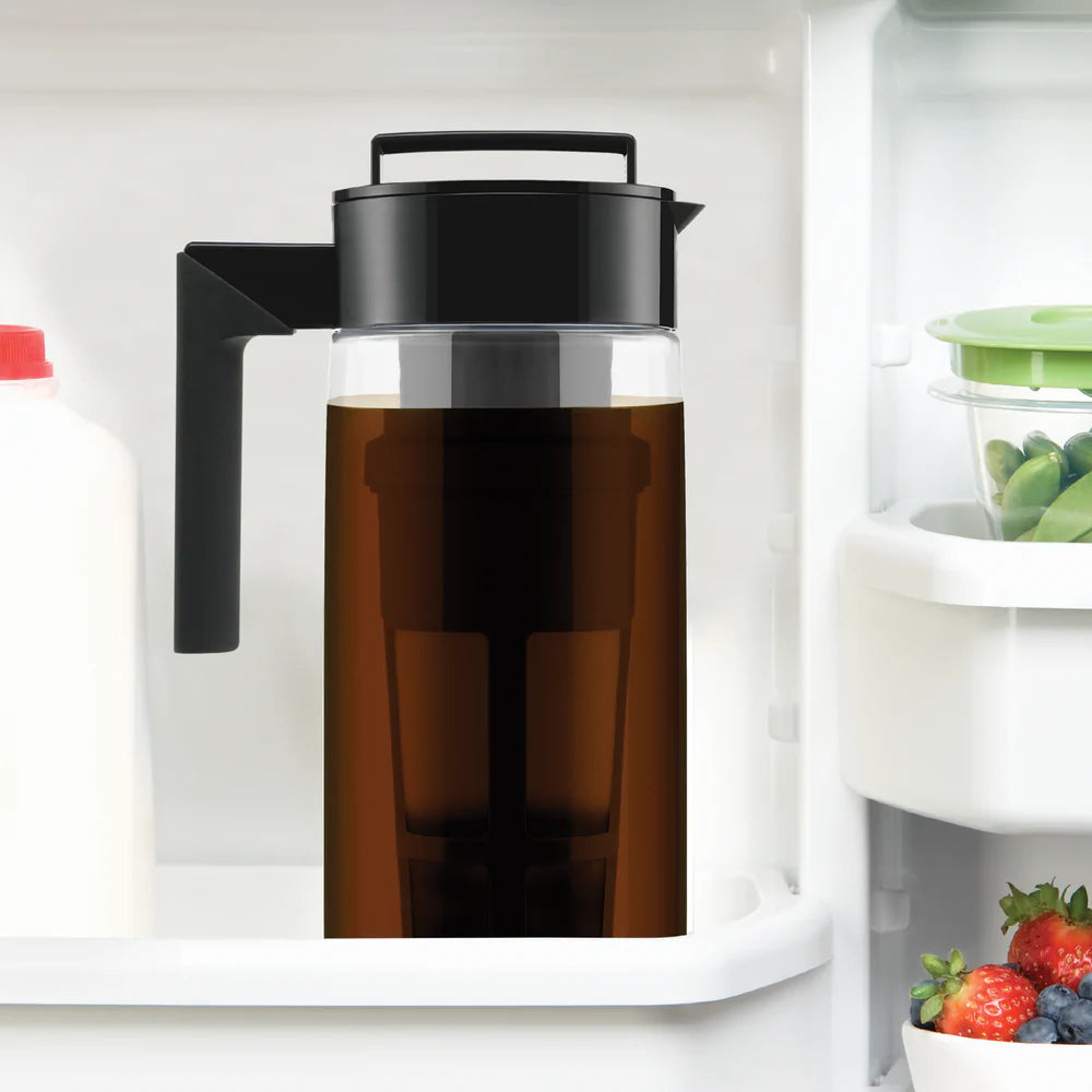 A sleek black and transparent pitcher filled with dark liquid sits in a refrigerator next to milk and fresh produce, ideal for organic teas.